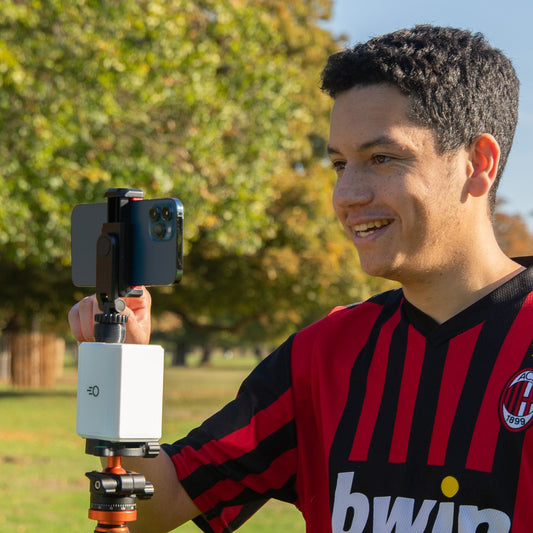 A man wearing a soccer jersey stands outdoors in a park holding a smartphone mounted on a Pitchwise Mount.
