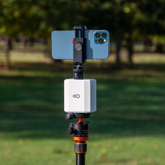 A Pitchwise Mount, mounted on a black tripod in an outdoor park setting with a smartphone attached to its top.